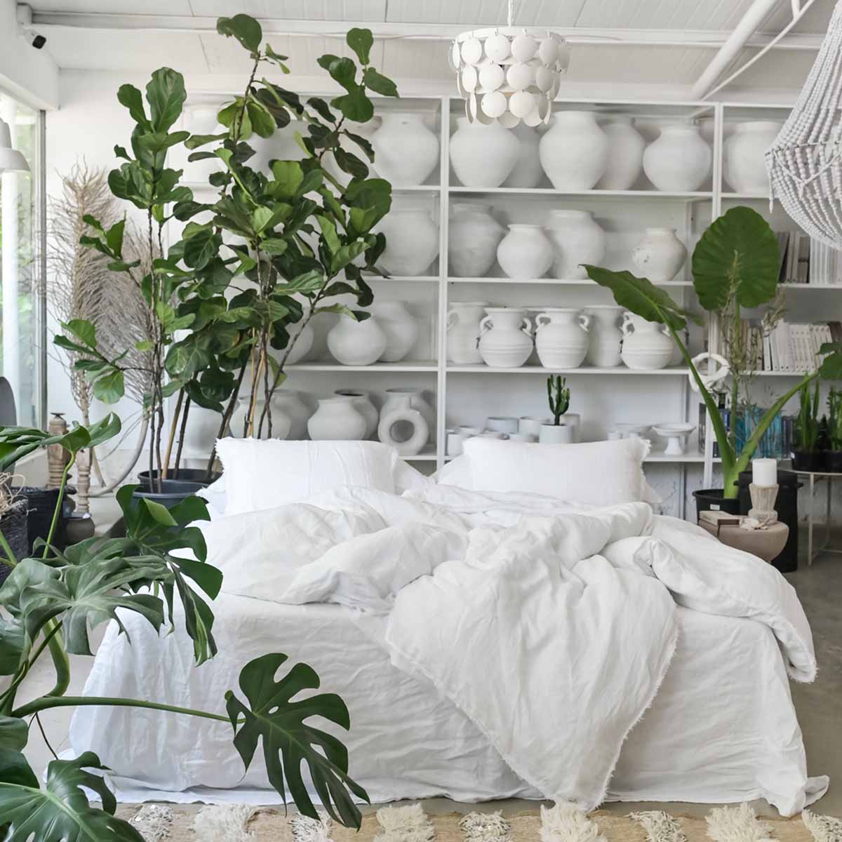 מיטה עטופה במצעי פשתן לבן של Fine Lab Home ניצבת מול מדפים עם אגרטלים וכלי חרס לבנים, מוקפת בצמחי בית ירוקים ושופעים בחדר מואר.
