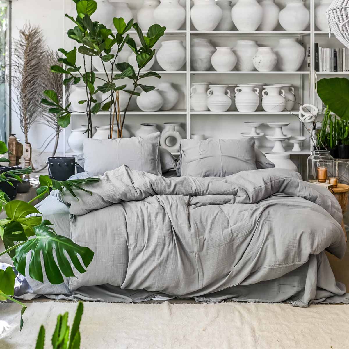 מיטה עם ציפה אפור טטרה מקומטת מבית Fine Lab Home ומצעים אפורים ניצבת מול מדפים לבנים עמוסים באגרטלי קרמיקה, מוקפים בצמחי בית ירוקים גדולים.