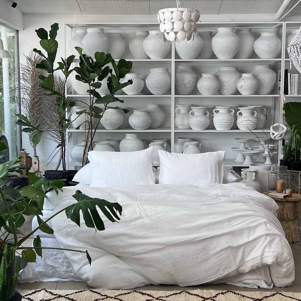 מיטה לבנה עם סט מצעים טטרה זוגי לבן – סדין 1.80 מבית Fine Lab Home ניצבת מול מדפים שעליהם אגרטלי קרמיקה לבנים גדולים, וצמחים ירוקים מקשטים את החדר.