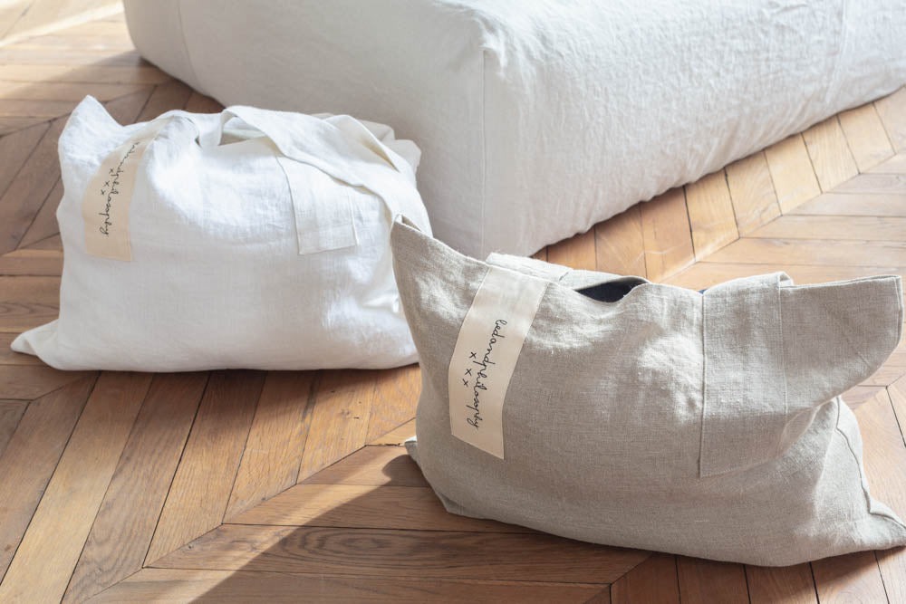 תיק פשתן של Fine Lab Home הוא תיק נשיאה מלבני בצבע לבן-שמנת, עם שתי ידיות רחבות ותווית קטנה בצבע בז' עם כיתוב בכתב יד.