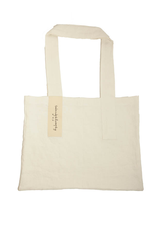 תיק פשתן של Fine Lab Home הוא תיק נשיאה מלבני בצבע לבן-שמנת, עם שתי ידיות רחבות ותווית קטנה בצבע בז' עם כיתוב בכתב יד.