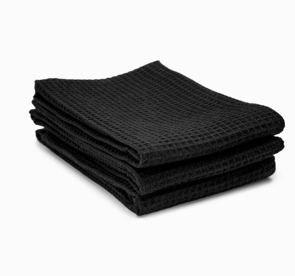 סט מגבות מטבח - צבע שחור 3 יח' מבית Fine Lab Home: שלוש מגבות פיקה שחורות עשויות כותנה 100%, מקופלות ומונחות אחת על גבי השנייה על רקע לבן.