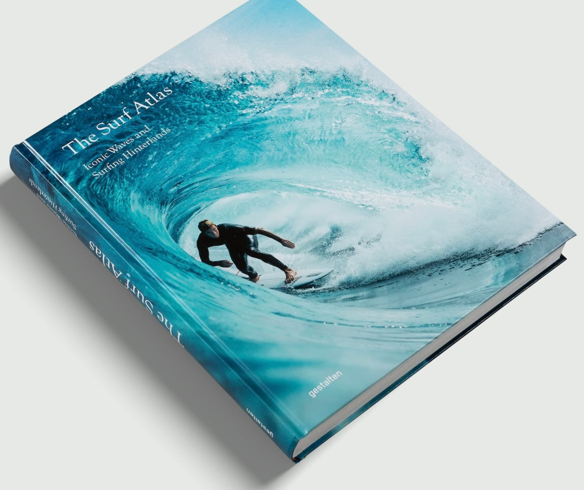 ספר העיצוב "The Surf Atlas" בכריכה קשה מבית Fine Lab Home כולל תמונת שער של גולש גלים בתוך גל כחול ענק.