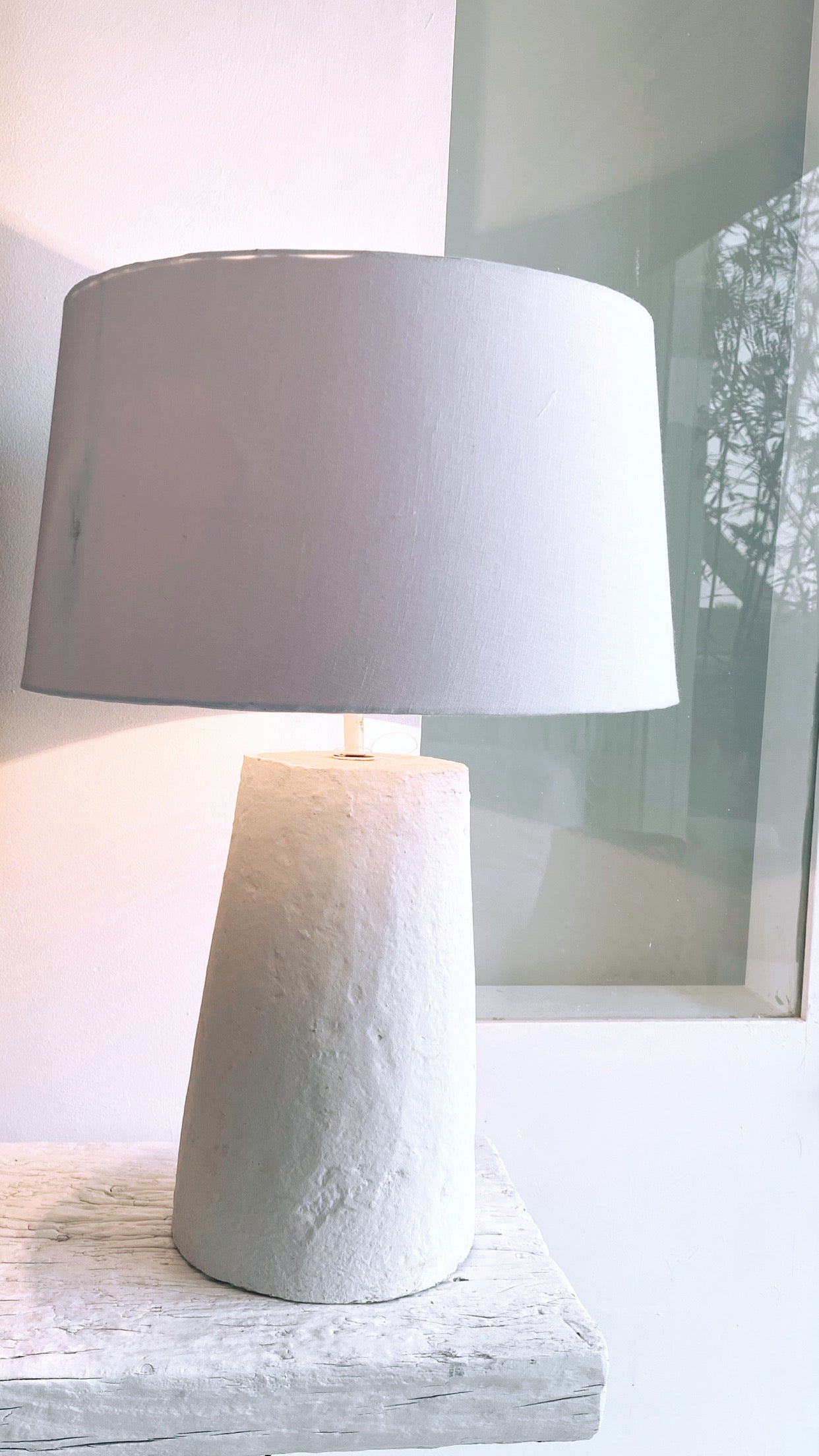 גוף תאורה ג'ואי מבית Fine Lab Home – מנורת שולחן לבנה עם בסיס גלילי מחוספס וצלון רחב בעיצוב מינימליסטי. כבל החשמל השחור מונח לצד גוף התאורה בעבודת יד.