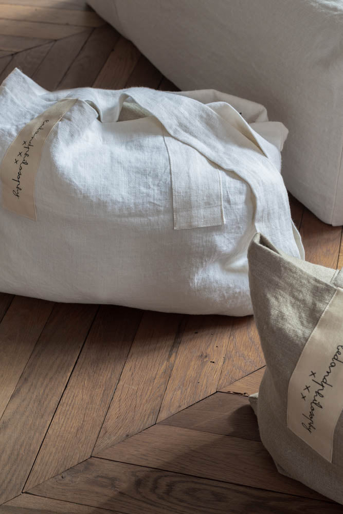 תיק פשתן של Fine Lab Home הוא תיק נשיאה מלבני בצבע לבן-שמנת, עם שתי ידיות רחבות ותווית קטנה בצבע בז' עם כיתוב בכתב יד.