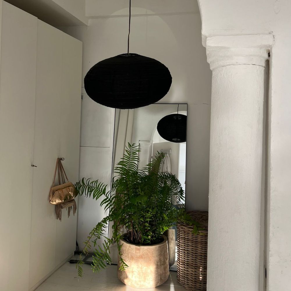 שרך בעציץ מונח על הרצפה לצד סל נצרים ומראה קיר בחדר בעל קירות לבנים, המואר על ידי אהיל תאורה בצורת כיפה מבית Fine Lab Home, עשוי פשתן שחור.