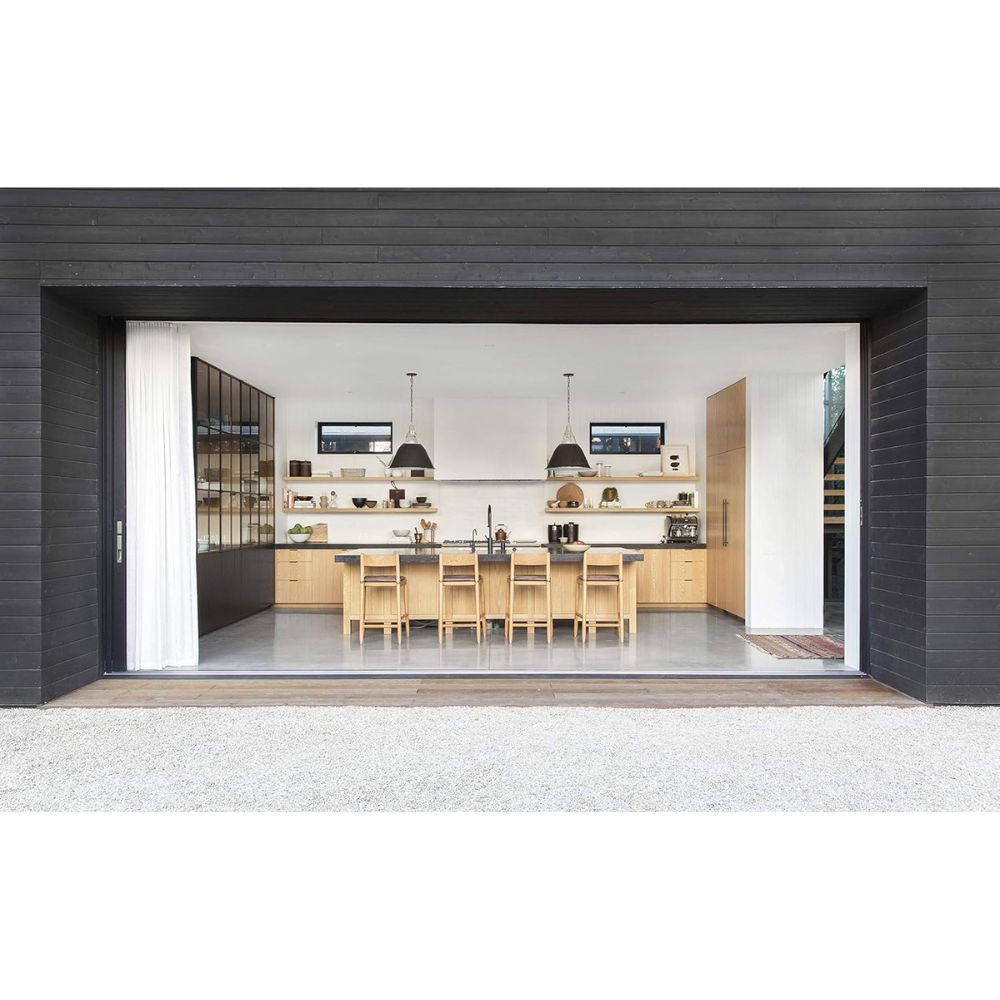 מבט פתוח על מטבח מודרני עם ארונות עץ בהירים, שולחן אוכל, מנורות תלייה ומדפים פתוחים — השראה מושלמת לחובבי עיצוב הבית ומעריצי אמבר לואיס או ספר העיצוב של Fine Lab Home שנוצר למען החיים.