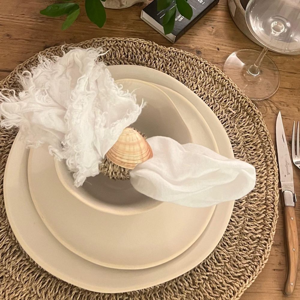 סידור שולחן עם סט צלחות como מבית Fine Lab Home — מפה ארוגה, צלחות וקערה בצבע שמנת בעבודת יד, מפיות עם טבעת צדף, סכו"ם וכוס יין על שולחן עץ.