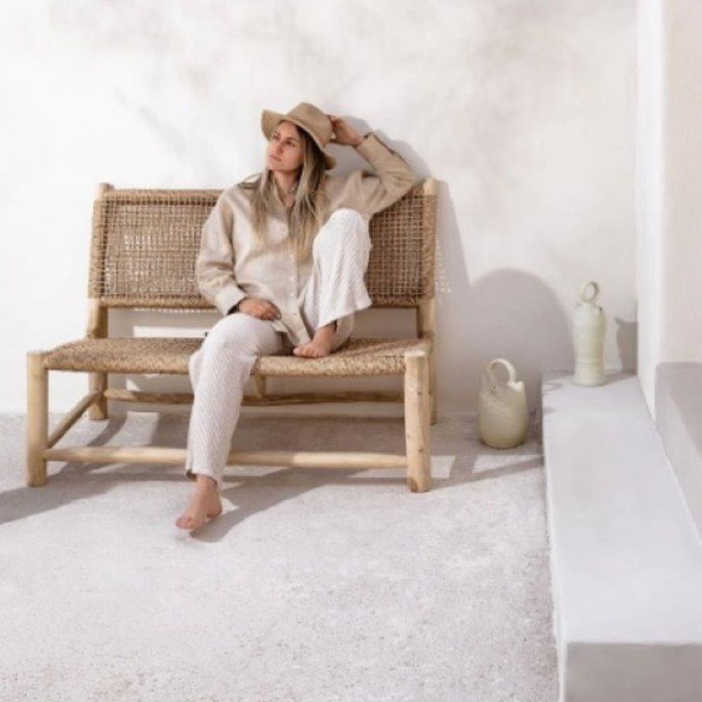 אישה בבגדים קלים וכובע יושבת יחפה על ספה מעץ טיק וראטן של Fine Lab Home על רקע קיר לבן, כששני כדים קרמיים מונחים על הרצפה לידה.