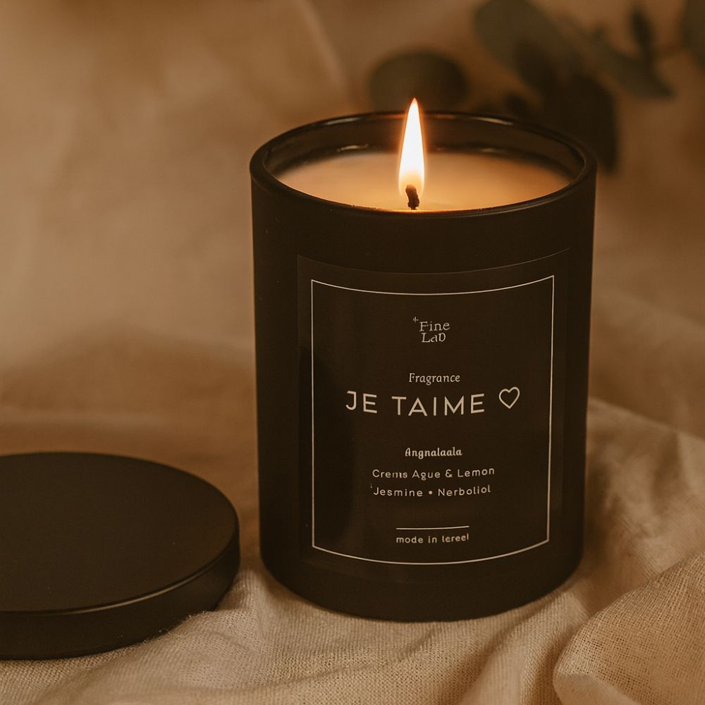 נר שחור דולק של Fine Lab Home, עם הכיתוב JE TAIME, מונח על בד עם מכסה לידו; הרקע המטושטש בעדינות כולל גוונים ניטרליים.