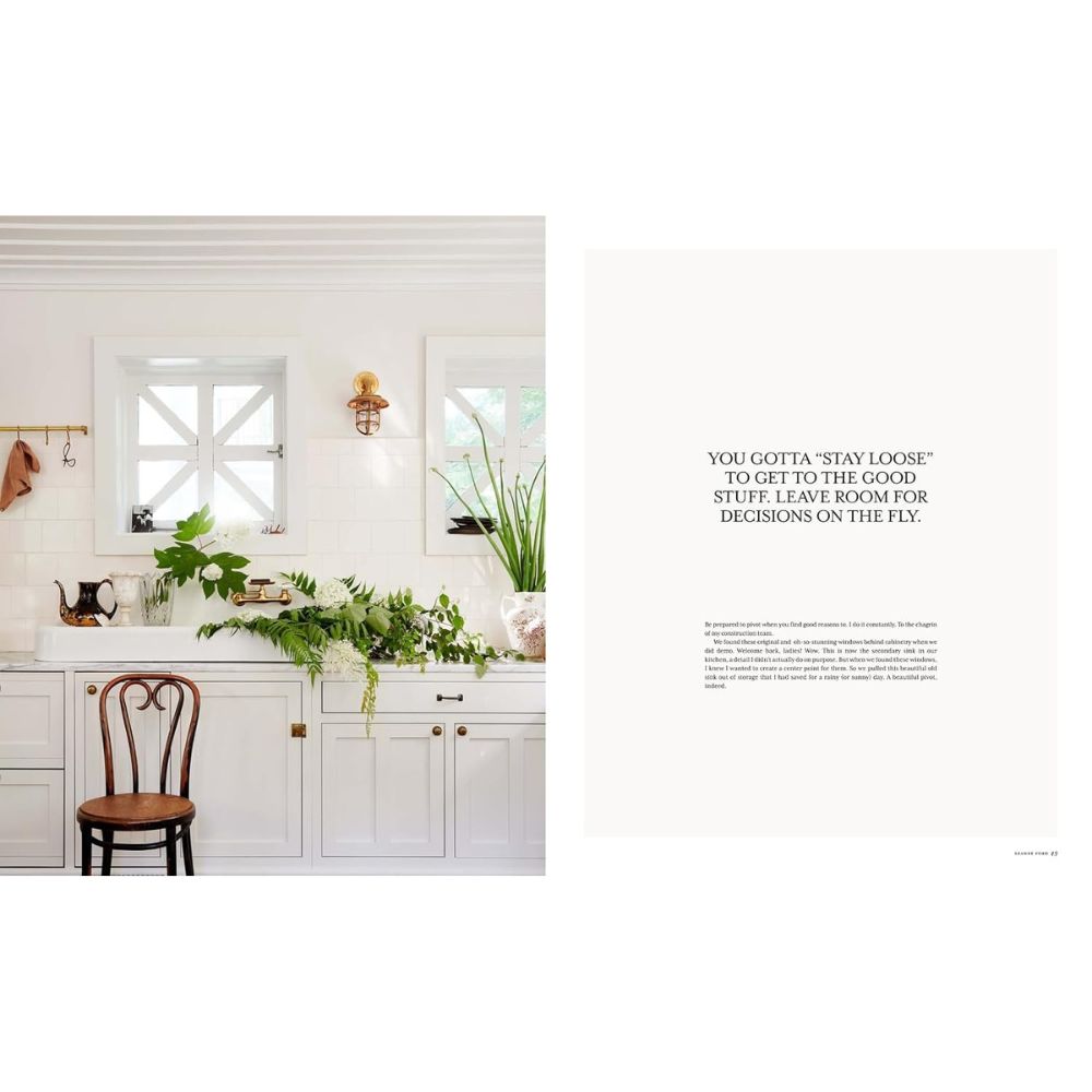 מטבח מואר עם ארונות לבנים, כיסא עץ וצמחים על השיש; לידם מונח ספר העיצוב The Slow Down של Fine Lab Home.