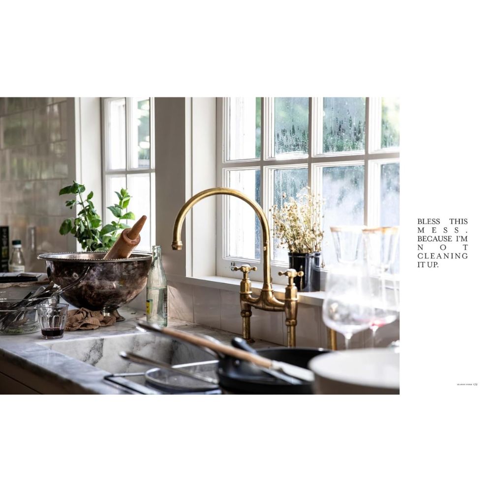 כיור מטבח עם כלים וקערות מלוכלכים ניצב מתחת לחלון עם צמחים; הטקסט אומר: "ברך את הבלגן הזה" — בהשראת ספר העיצוב The Slow Down מבית Fine Lab Home.