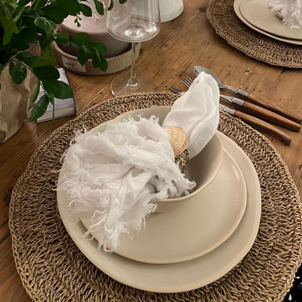 סט צלחות como מבית Fine Lab Home בצבע בז', מפה ארוגה, סכו"ם, כוס יין ומפית לבנה מעוטרת בצדף יוצרים עיצוב מינימליסטי על שולחן עץ.