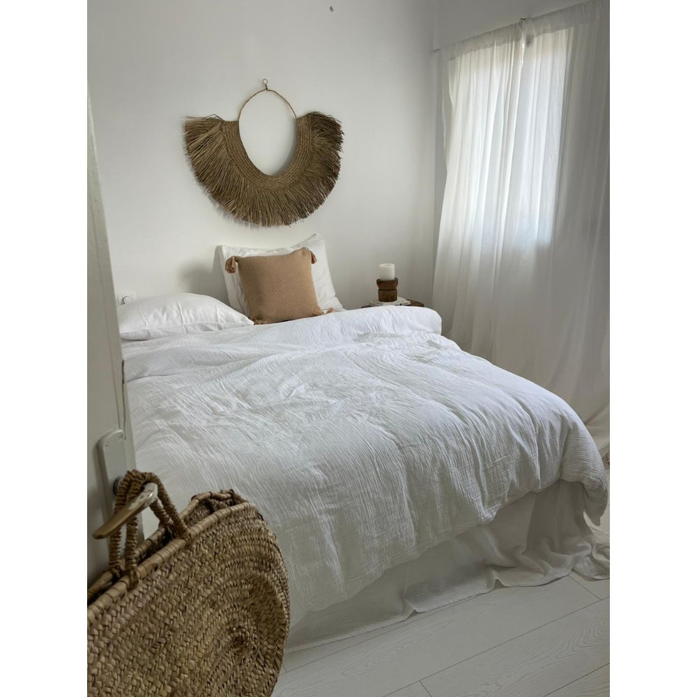 חדר שינה מינימליסטי עם מיטה מסודרת, הכוללת סט מצעים טטרה זוגי לבן – סדין 1.60 מבית Fine Lab Home, כרית בצבע בז', יצירת אמנות ארוגה על הקיר מעל ראש המיטה, וחלונות המעוטרים בווילונות לבנים שקופים.