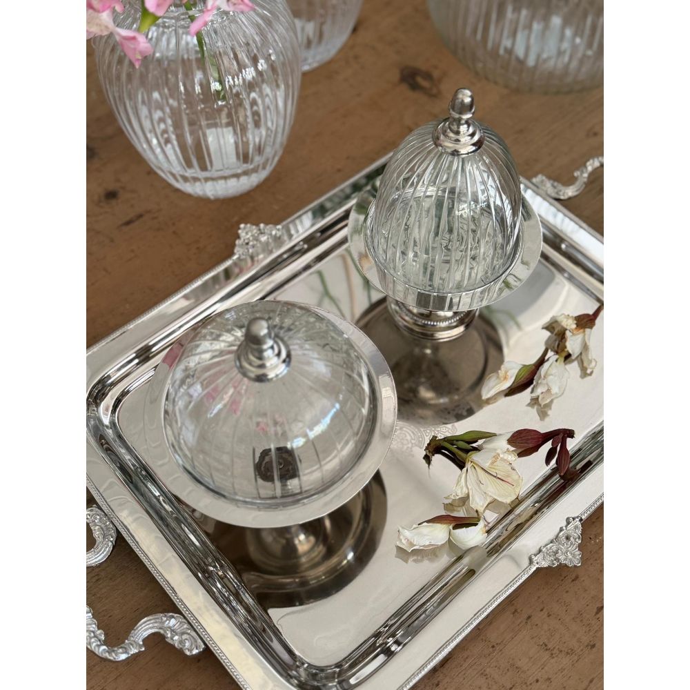 The Fine Lab Home פעמון זכוכית וכסף אלגנטי- שלושה גדלים, המציג כיפות זכוכית עם בסיסים ומכסים מכסף, מוצג על מגש כסף רפלקטיבי עם עלי כותרת לבנים מפוזרים ואגרטלי זכוכית ברקע.