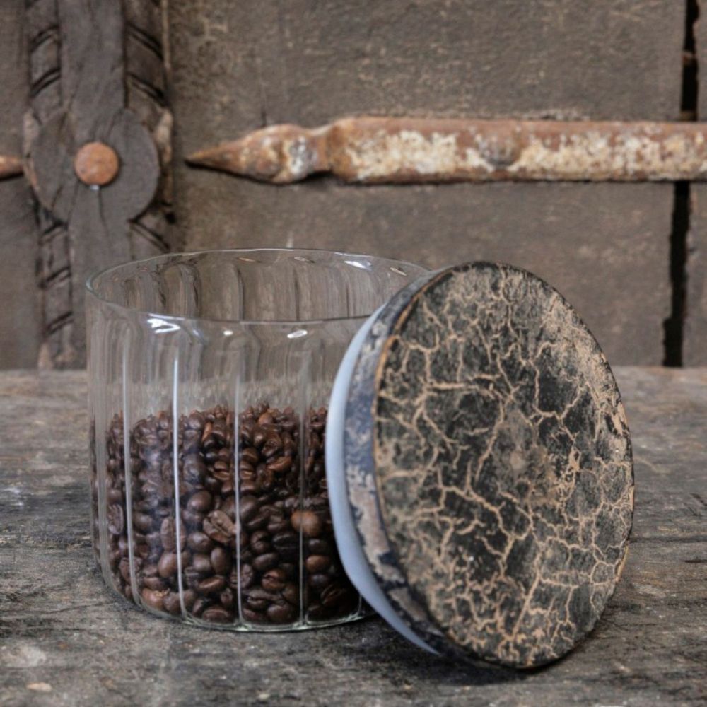 צנצנת זכוכית מפוספסת עם מכסה עץ מבית Fine Lab Home, מלאה בפולי קפה, מונחת על משטח מחוספס לצד המכסה שלה.