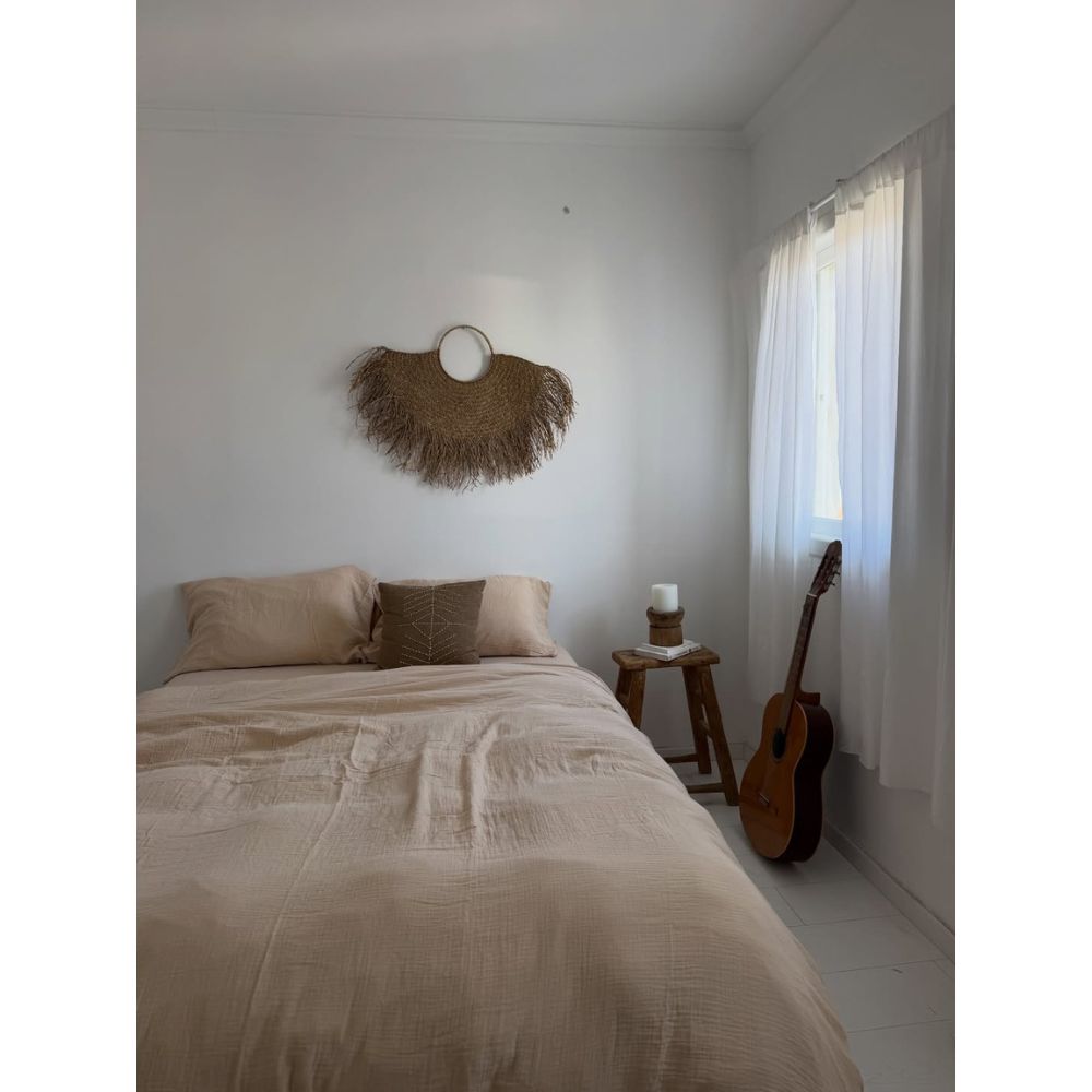 מיטה מסודרת עם סט מצעים טטרה זוגי בצבע חול של Fine Lab Home – סדין 1.80 מטר בצבע בז', שולחן צד מעץ עם נר, גיטרה צמודה לקיר, ותמונה ארוגה התלויה מעל, באור רך.