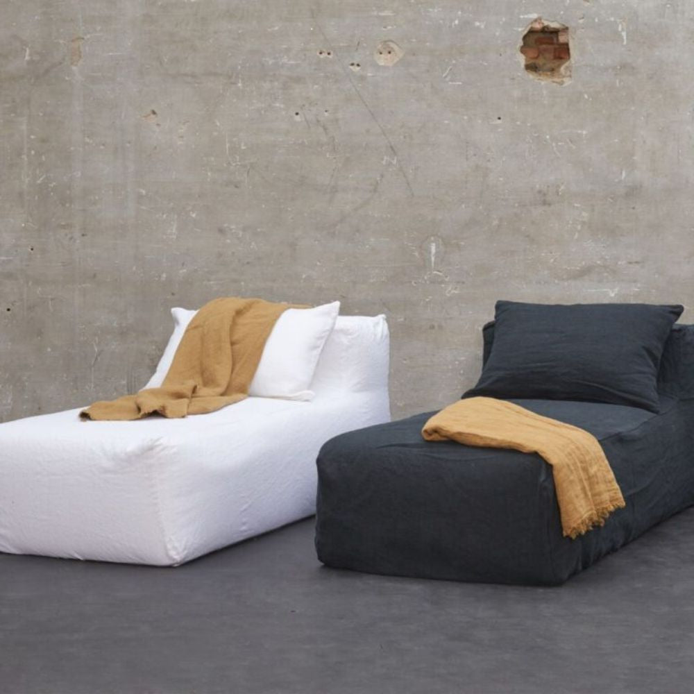 שתי מיטות סלון של Fine Lab Home, עשויות 100% פשתן – האחת עם מצעים לבנים והשנייה באפור כהה – כשעל כל אחת מהן מונחת שמיכת פשתן בצבע צהוב חרדל; הן ניצבות מול קיר בטון חשוף עם כתם קטן של לבנים חשופות.