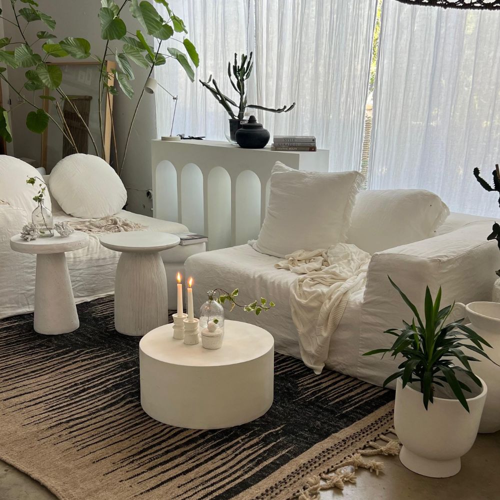 סלון מודרני עם ריהוט לבן, עציצים, סטול לבן של Fine Lab Home ליד נרות על שולחן הקפה, ווילונות שקופים המאפשרים לאור טבעי להיכנס.