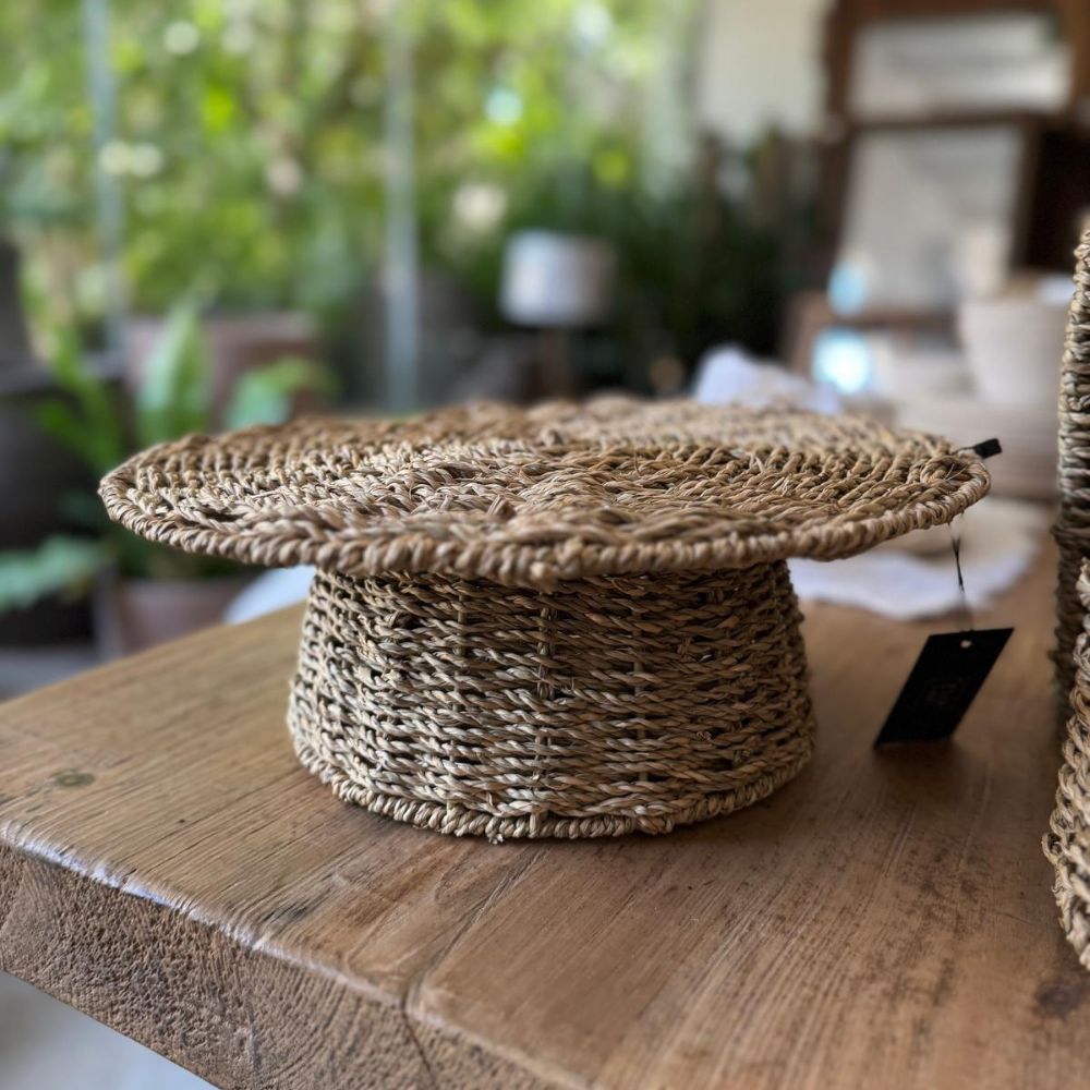 המגש העגול על רגל מחבל טבעי של Fine Lab Home הוא מגש עגול, ארוג, עם חלק עליון רחב ושטוח ובסיס צר, עשוי מסיבים טבעיים ומוצג על רקע לבן חלק.