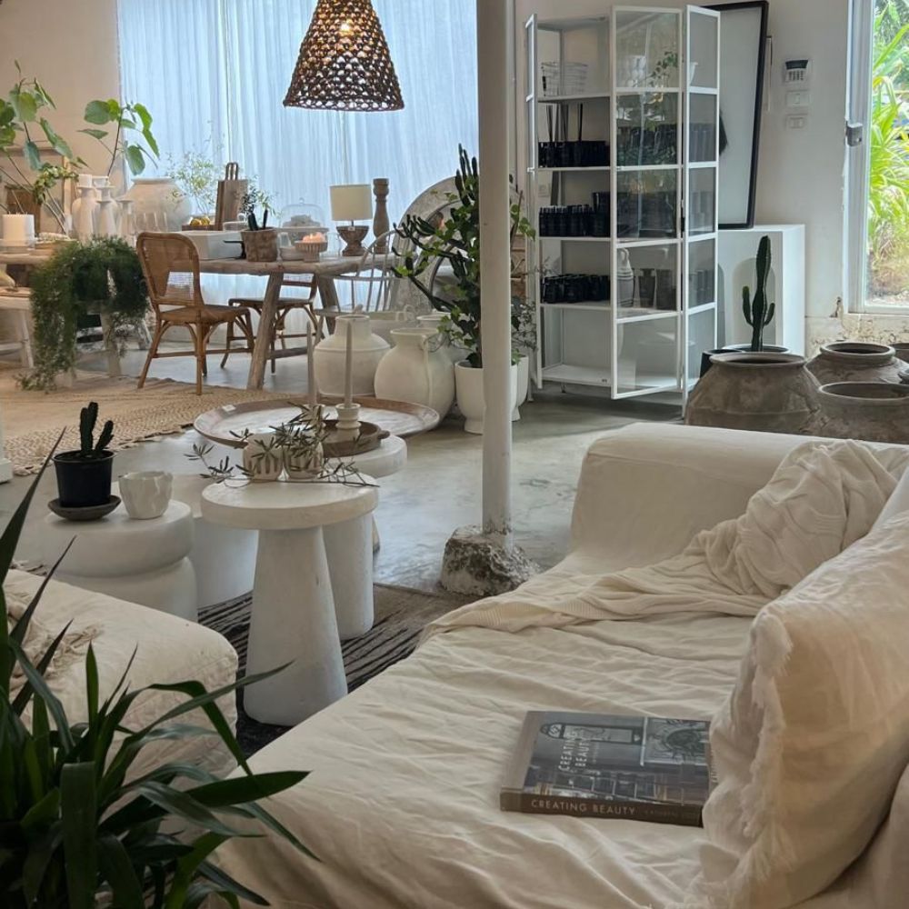 סלון נעים ומואר עם ספה לבנה, שולחנות עגולים עם צמחים, סטול לבן מבית Fine Lab Home, מדף ספרים, שולחן אוכל, צמחים שונים ומגזין בשם Creating Beauty על הספה.
