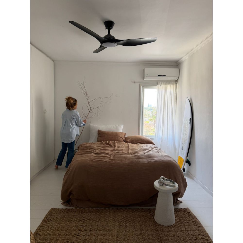 אדם מסדר ענפים ליד המיטה עם סט מצעים טטרה זוגי חום - סדין 1.60 של Fine Lab Home בחדר שינה מינימליסטי; גלשן נשען על הקיר ליד חלון מואר באור השמש.