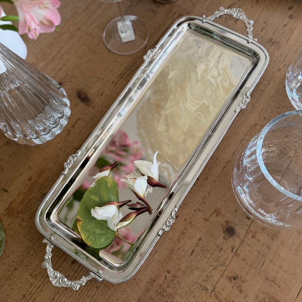The Fine Lab Home מגש כסף אלגנטי עם ידיות- שני גדלים משקף פרחים ורודים ולבנים, מוקפים בכלי זכוכית על שולחן עץ למראה אלגנטי.