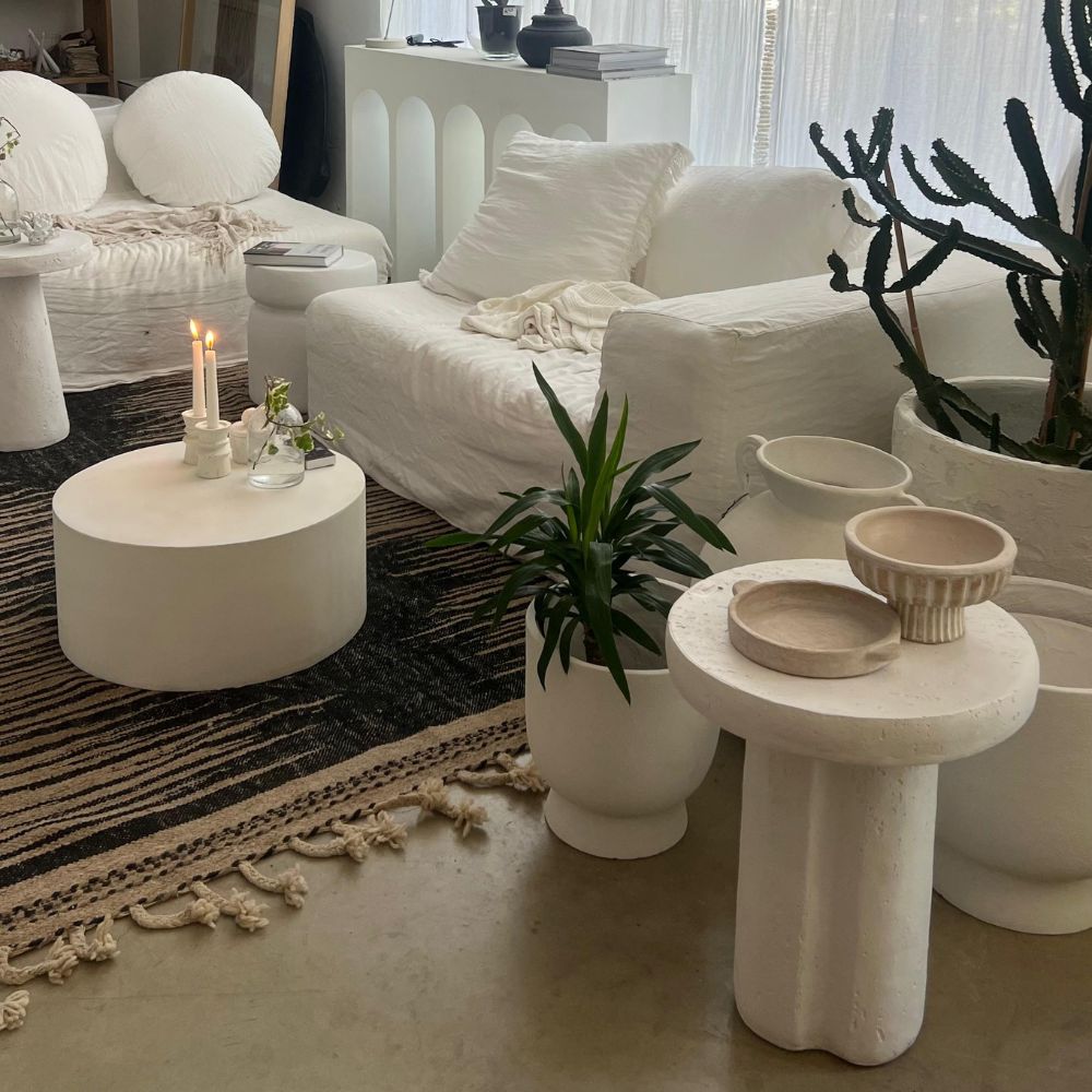 סלון מודרני עם סטול לבן- בסיס גליל של Fine Lab Home, ריהוט לבן, עציצים, שטיח פסים ונרות דולקים על שולחן קפה עגול.