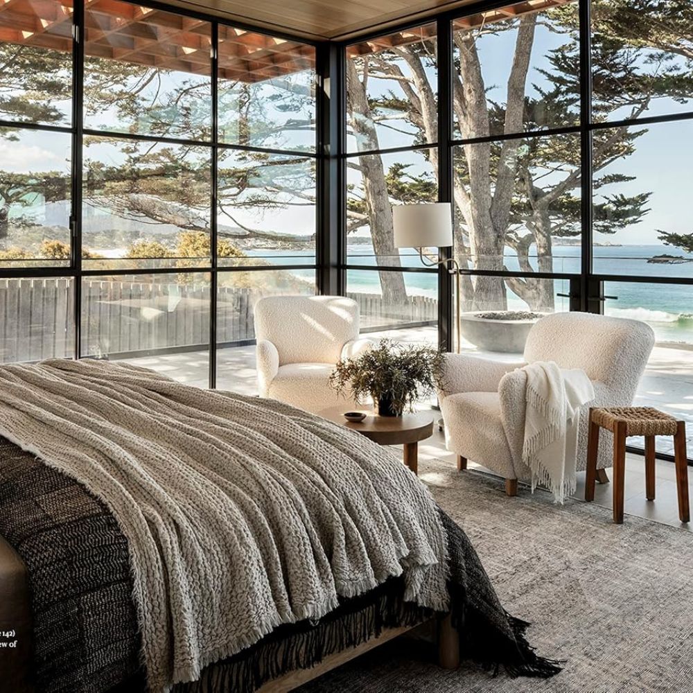 חדר שינה מודרני עם חלונות גדולים, נוף לים, שני כורסאות לבנות, שולחן קטן ומיטה עם שמיכות ניטרליות ומרקמות – מושלם לחובבי עיצוב פנים או לקוראי ספר עיצוב. קראו לו בית מאת Fine Lab Home.