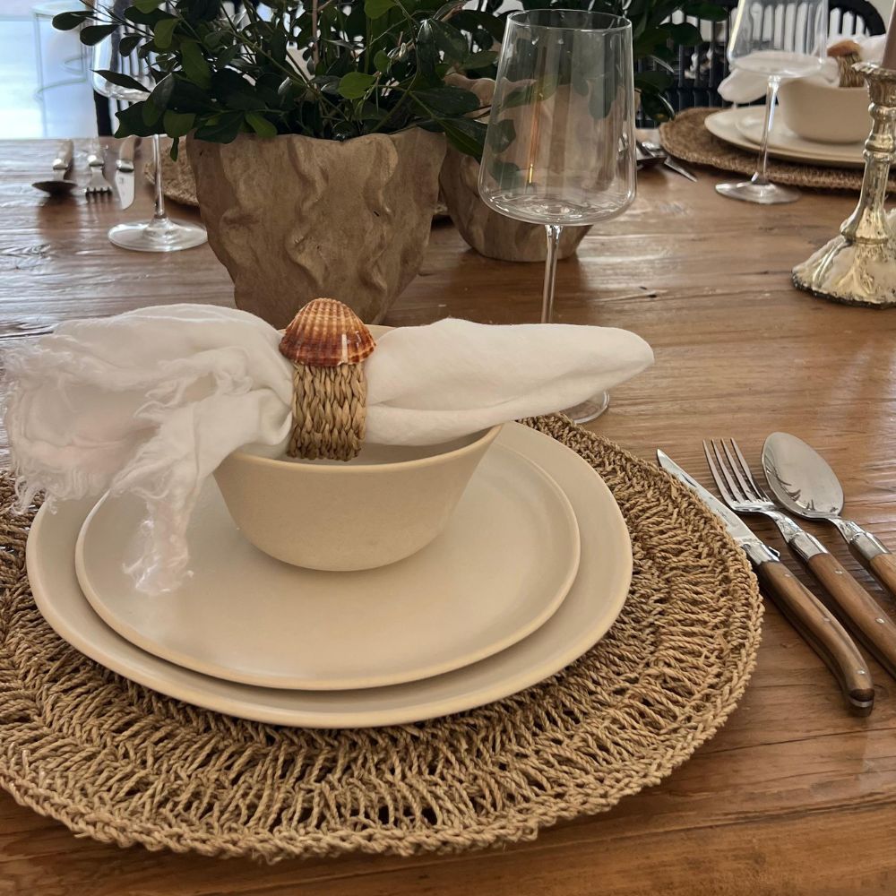 שולחן ערוך עם מפה ארוגה, סט צלחות Fine Lab Home como, קערה, מפיות לבנות בטבעת צדף, סכו"ם, כוסות, ומרכז שולחן עלים על שולחן עץ בעיצוב מינימליסטי.