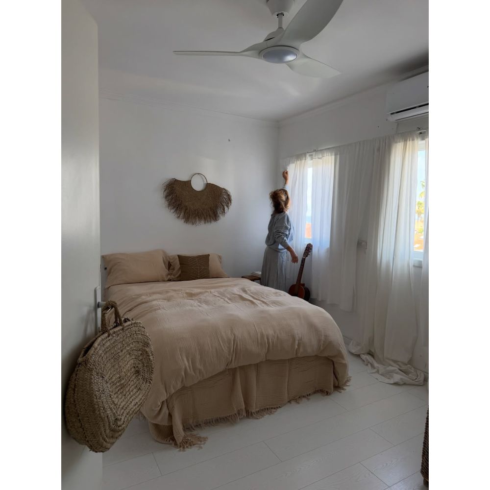 אישה עומדת ליד חלון בחדר שינה מינימליסטי, שבו ניתן לראות את סט המצעים טטרה זוגי חול של Fine Lab Home – סדין 1.80, קישוט קיר ארוג וגיטרה המונחת בסמוך.