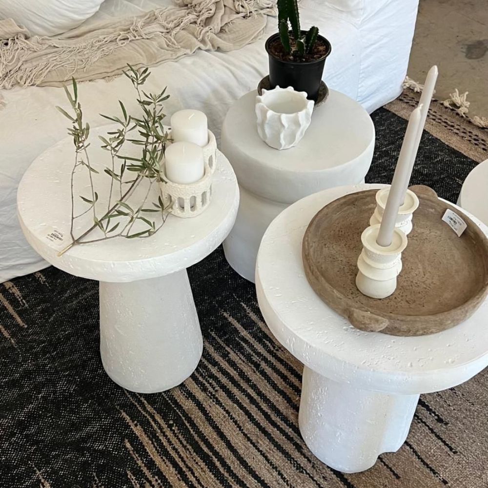 שלושה שולחנות צד לבנים, כולל שולחן צד לבן, שולחן צד מינימליסטי וסטול לבן מבית Fine Lab Home, מעוטרים בנרות, עציץ, ענף וכוס קרמיקה על שטיח שחור-בז' לצד ספה לבנה.