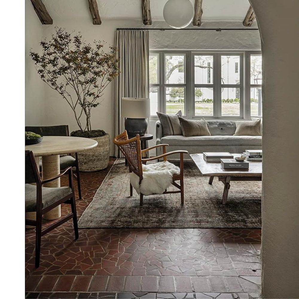 סלון המעוצב בסגנון עיצוב פנים בהשראת אמבר לואיס, עם ספר העיצוב Call It Home מבית Fine Lab Home על שולחן הקפה, ספה אפורה, שולחן קפה מעץ, שולחן אוכל עגול, קורות חשופות, חלונות גדולים ועציצים.