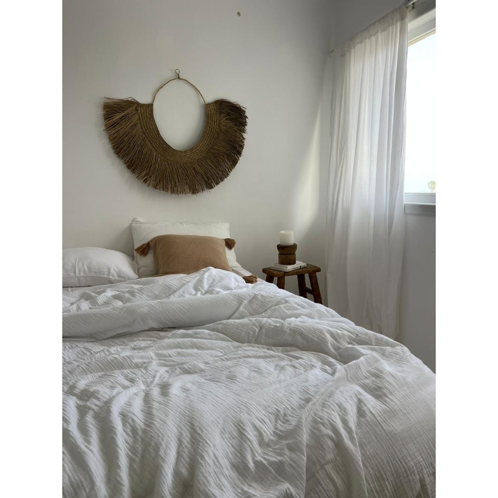 מיטה מסודרת עם סט מצעים טטרה זוגי לבן של Fine Lab Home – סדין 1.60, כרית בצבע חום בהיר, שרפרף עץ עם נר, וילונות שקופים, וקישוט קיר ארוג התלוי מעל ראש המיטה.