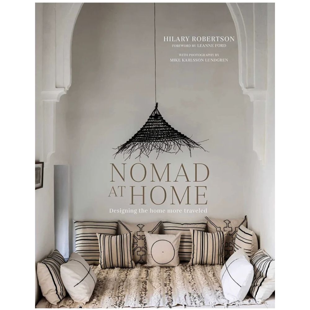 על עטיפת הספר עיצוב nomad at home מאת Fine Lab Home מוצג ספסל מרופד עם כריות מעוטרות ותליון תאורה ארוג בתוך גומחה מקושתת, המוסיפים את הנגיעה המושלמת של עיצוב נוודי.