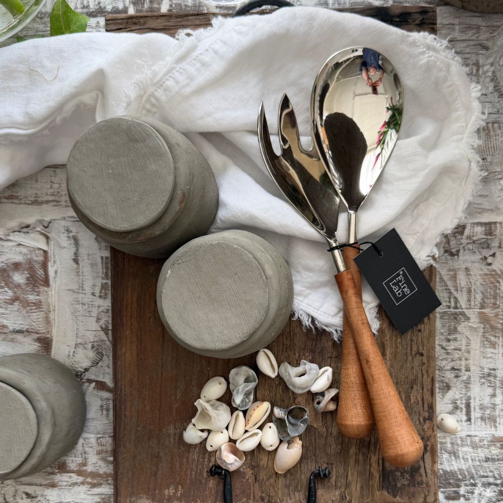 שני צנצנות קרמיקה אפורות, כף ומזלג לסלט מעץ וניקל של Fine Lab Home עם ידיות עץ, ומבחר צדפים מסודרים על משטח עץ המכוסה במפה לבנה.