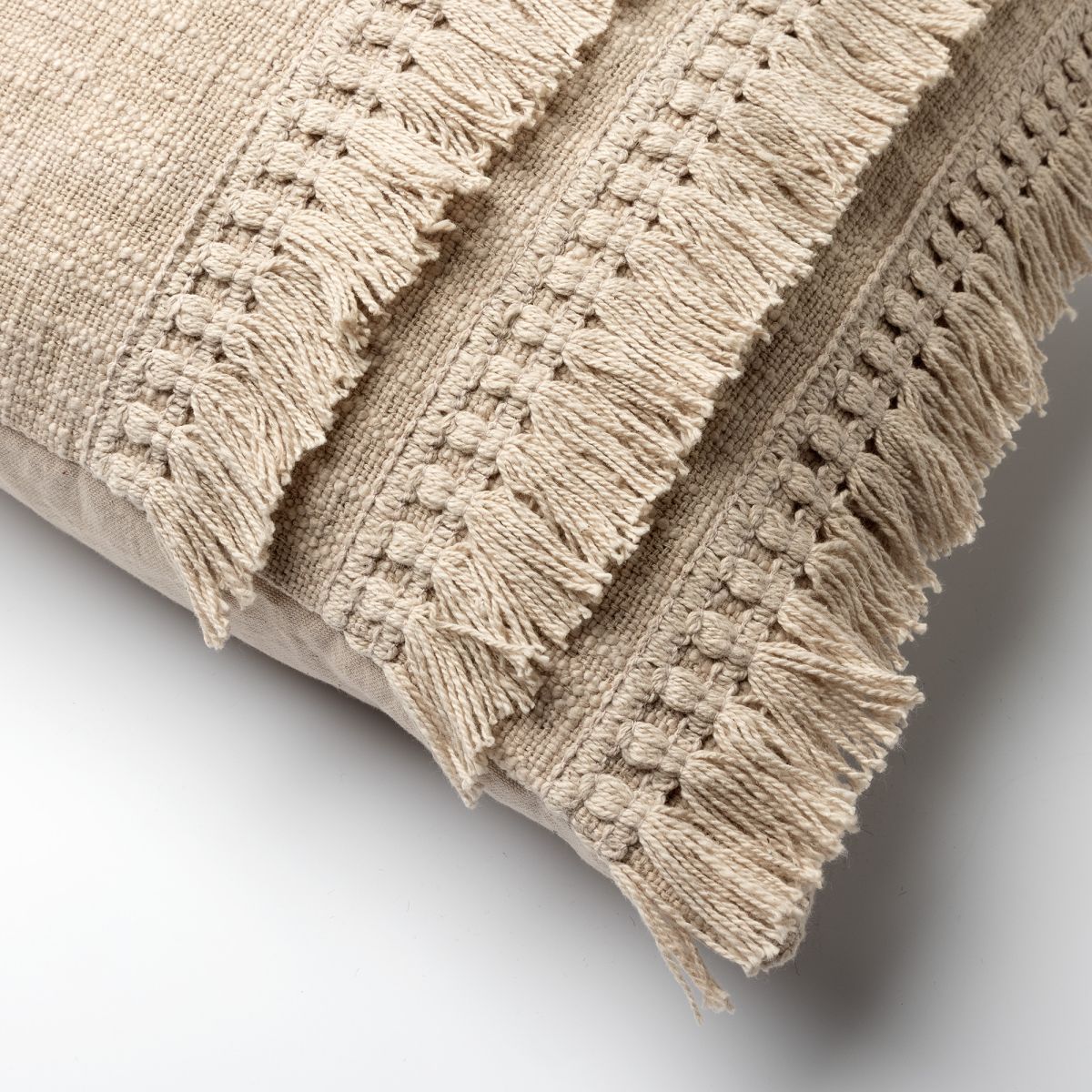 תקריב של כרית הנוי המלבנית "אבן" מבית Fine Lab Home, בצבע בז' עם דוגמאות מרקמיות ושוליים עם ציציות, המונחת על משטח לבן חלק.