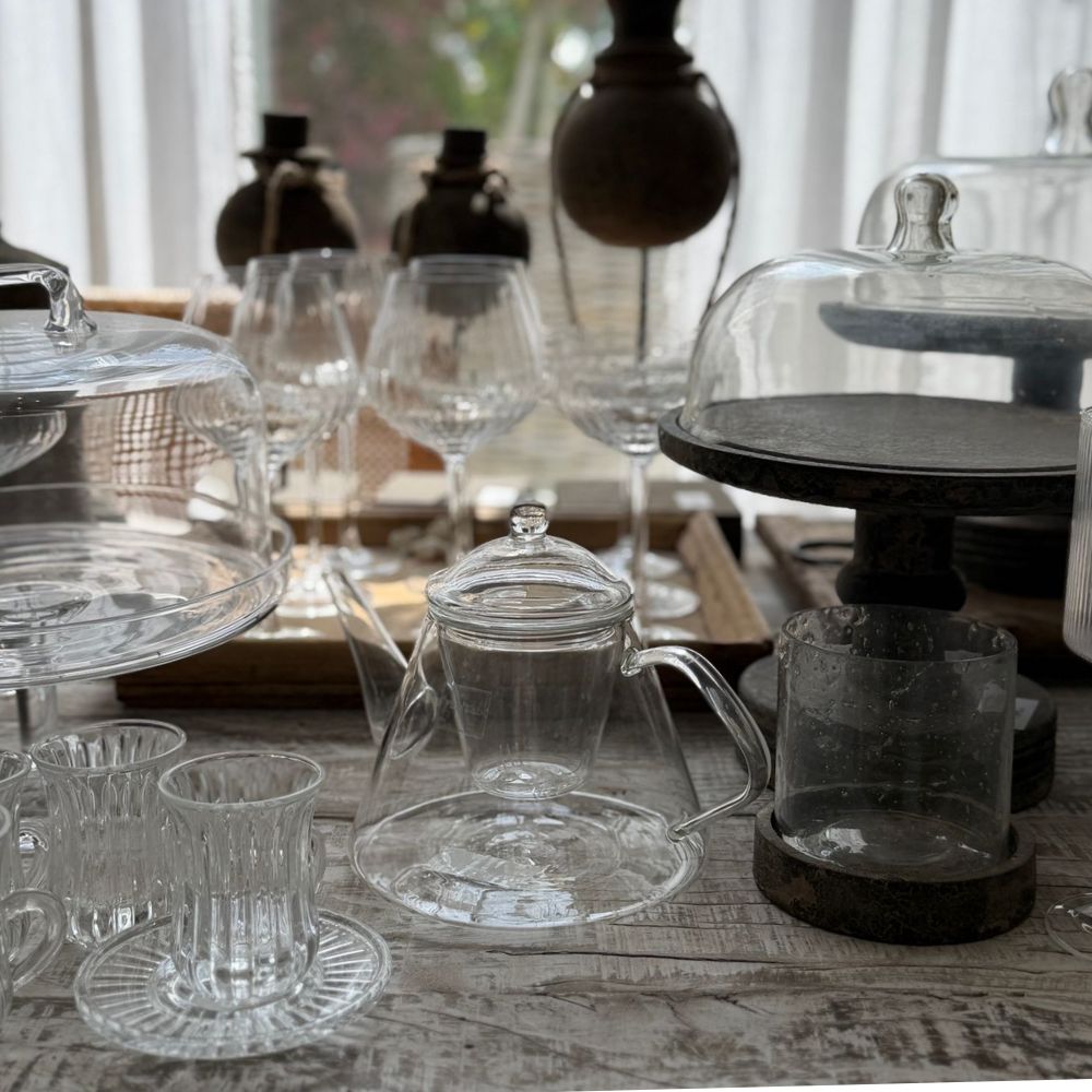 מגוון כלי זכוכית שקופים, בהם קנקן התה "Fine Lab Home", כוסות שתייה, כוסות יין ומעמדי עוגה, המוצגים על שולחן עץ בסגנון כפרי.