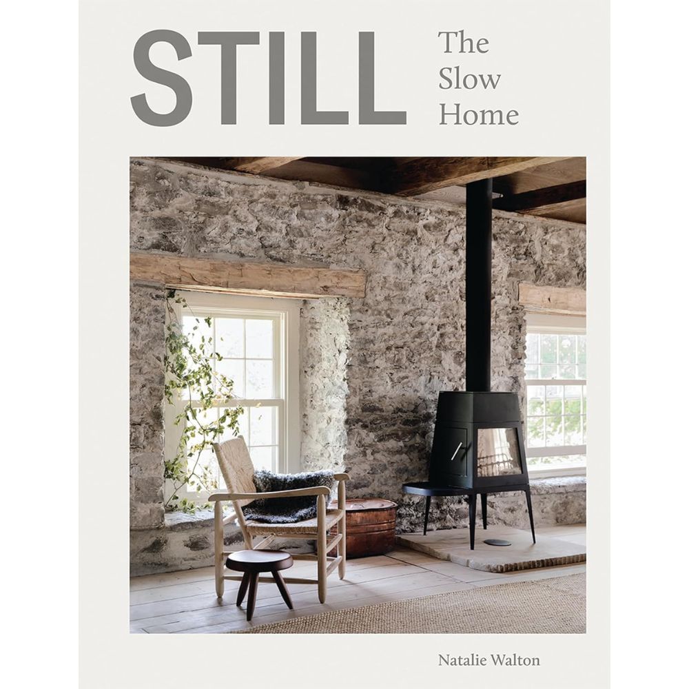 כריכת הספר "ספר עיצוב still the slow home" מאת Fine Lab Home, המציגה חדר כפרי עם קירות אבן, חלונות גדולים, תנור שחור וריהוט עץ מינימליסטי — דוגמה מעוררת השראה לעיצוב איטי ולחיים שלווים.