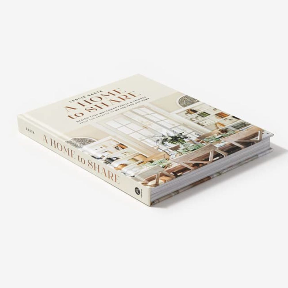 ספר העיצוב "A Home to Share" מבית Fine Lab Home מונח סגור על משטח לבן, וכעל הכריכה מופיע חדר מגורים מואר ומעוצב — השראה מושלמת לכל מי שאוהב לארח ועיצוב פנים.