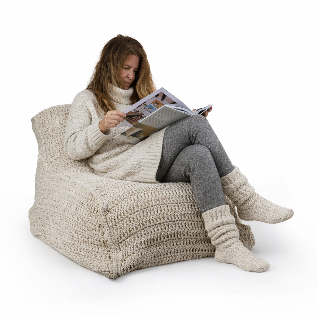 אדם לבוש בבגדי סריג נעימים יושב על פוף ישיבה-מקרמה של Fine Lab Home, קורא מגזין על רקע לבן, ומוסיף נופך של סגנון בוהו-שיק לסצנה.