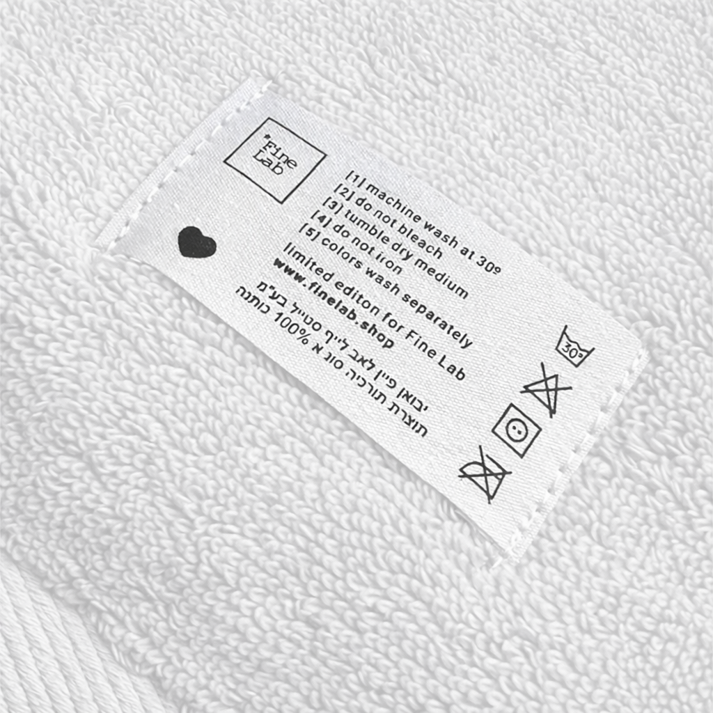 תקריב של תווית הטיפול בבד על זוג מגבות פנים/ידיים - צבע לבן מבית Fine Lab Home, המציגה הוראות כביסה באנגלית ובעברית. אידיאלי למי שמחפש מגבות כותנה או מגבות יוקרתיות.