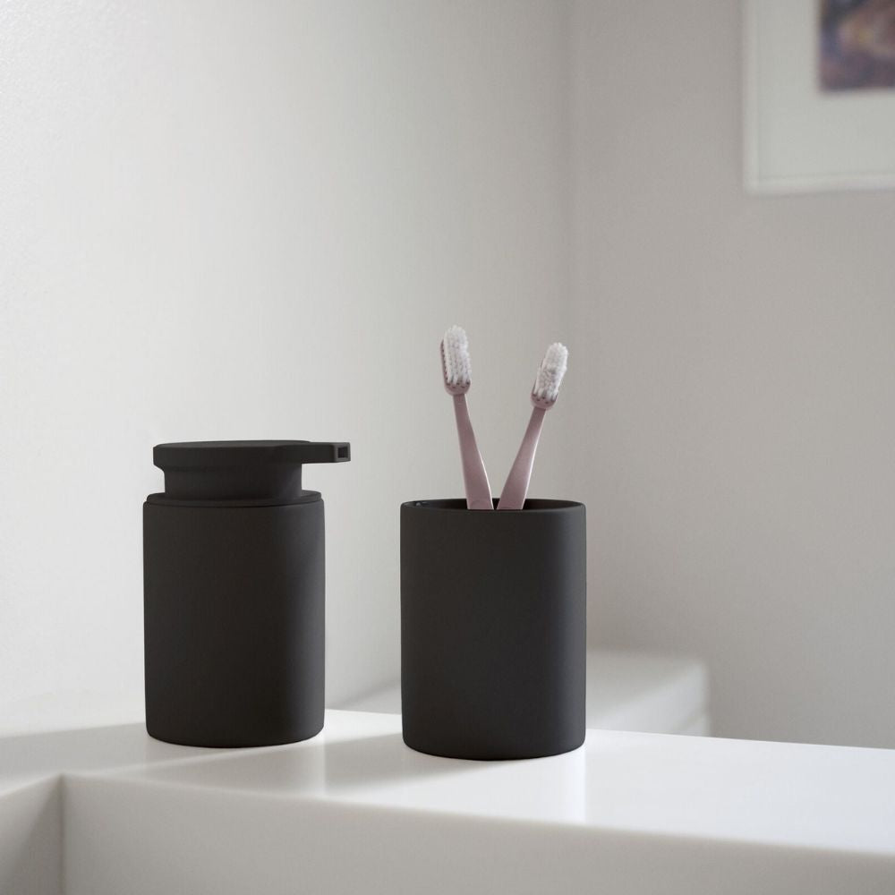 מתקן לסבון נוזלי בצבע שחור של Fine Lab Home וכוס תואמת עם שתי מברשות שיניים ורודות מונחים על משטח לבן בחדר האמבטיה.
