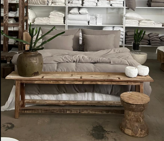 חדר מינימליסטי כולל מיטה מסודרת עם ציפית של Fine Lab Home - אפור סטון, מצעים בצבע בז', ספסל עץ מעוטר בכלי חרס וצמחים, וכן מדפים עמוסים במגבות ומצעים.