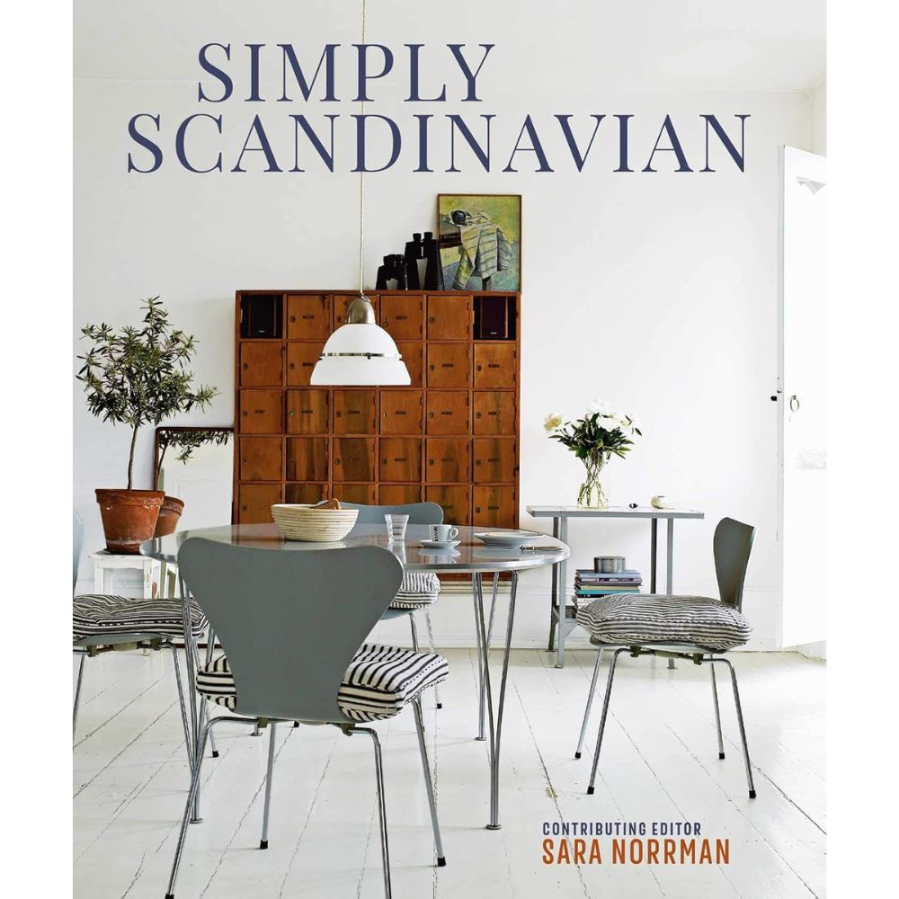 על כריכת ספר העיצוב "Simply Scandinavian" מבית Fine Lab Home מוצגת פינת אוכל מינימליסטית עם כיסאות אפורים, שולחן עגול, מנורה תלויה וארון עץ על רקע קיר לבן — המשקפים באופן מושלם את עקרונות העיצוב הסקנדינבי (Scandinavian design) ואת האלגנטיות המאופקת שבו.