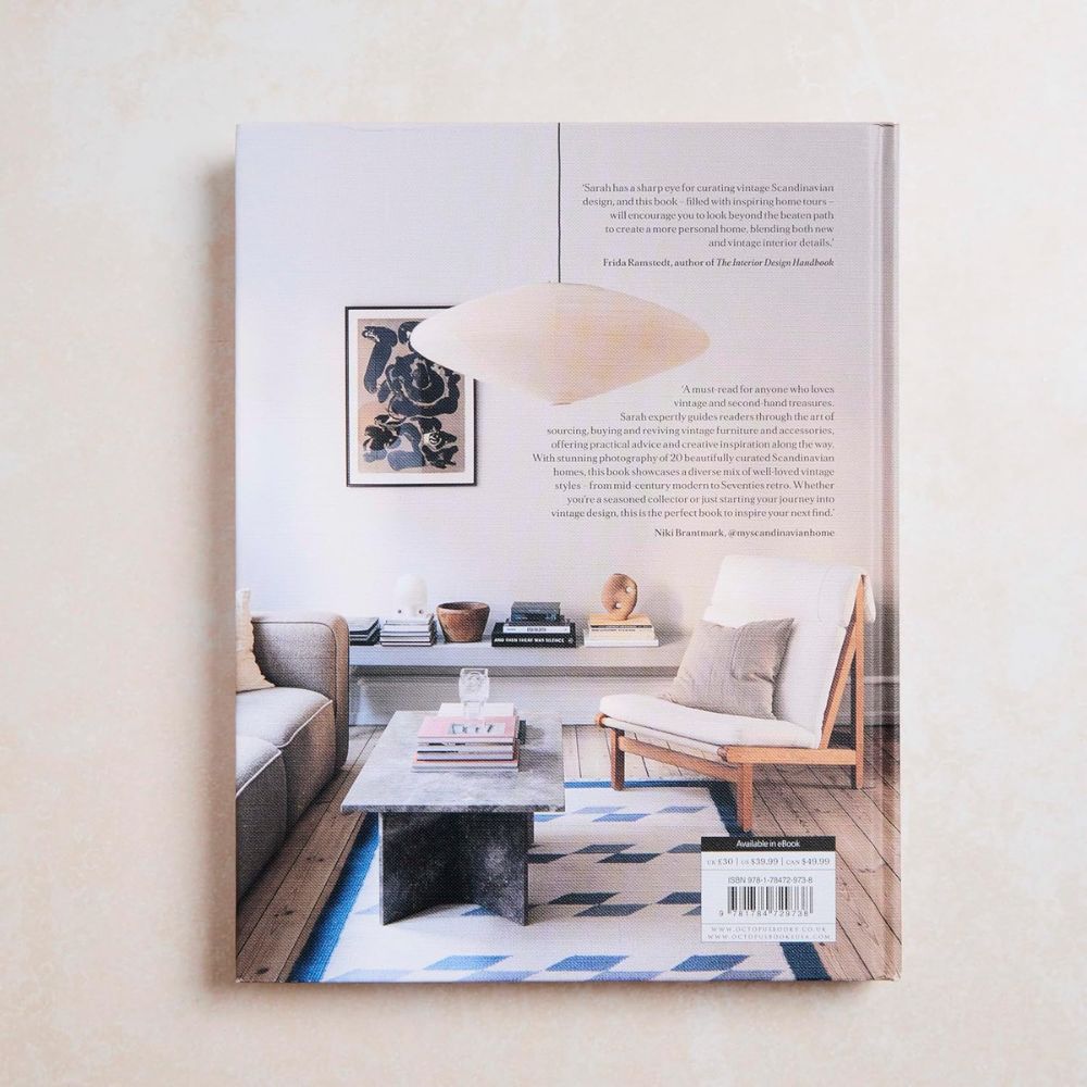 על הכריכה האחורית של הספר "The Vintage Way" מבית Fine Lab Home מוצג סלון מינימליסטי בסגנון סקנדינבי — כיסא עץ, ספה, שולחן קפה, תמונות קיר ומדפים עם ספרים — המהווה השראה לעיצוב פנים.