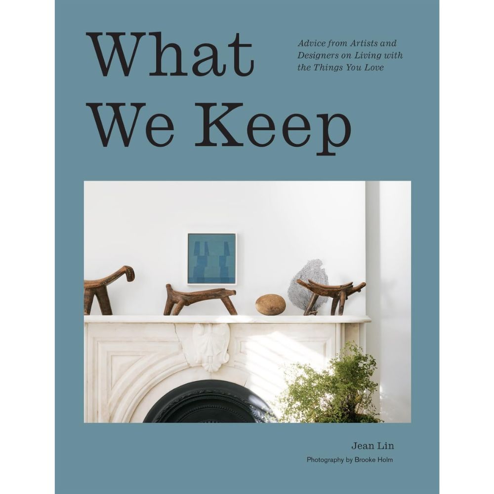 עטיפת הספר "What We Keep" מבית Fine Lab Home, המציגה מדף אח לבן מעוצב, המעוטר באביזרי נוי ובחפצי אמנות שנבחרו בקפידה על רקע כחול-אפור, המגלמת באופן יפהפה את רוח ספר העיצוב והעיצוב הפנים המודרני.
