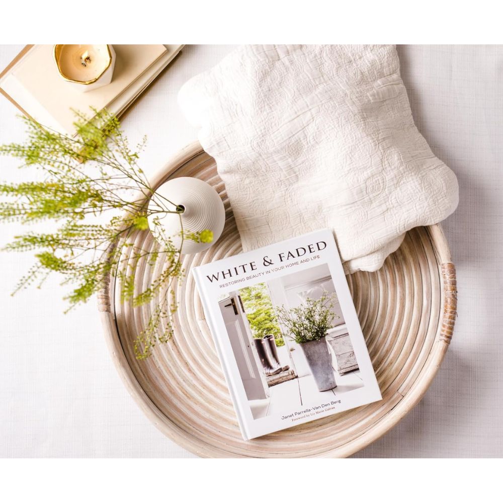 מגש קש עגול בסגנון כפרי נושא עליו מפה לבנה ומרקמת, את ספר העיצוב "White and Faded" מבית Fine Lab Home, אגרטל קטן עם צמחייה, ונר זהוב על משטח לבן.