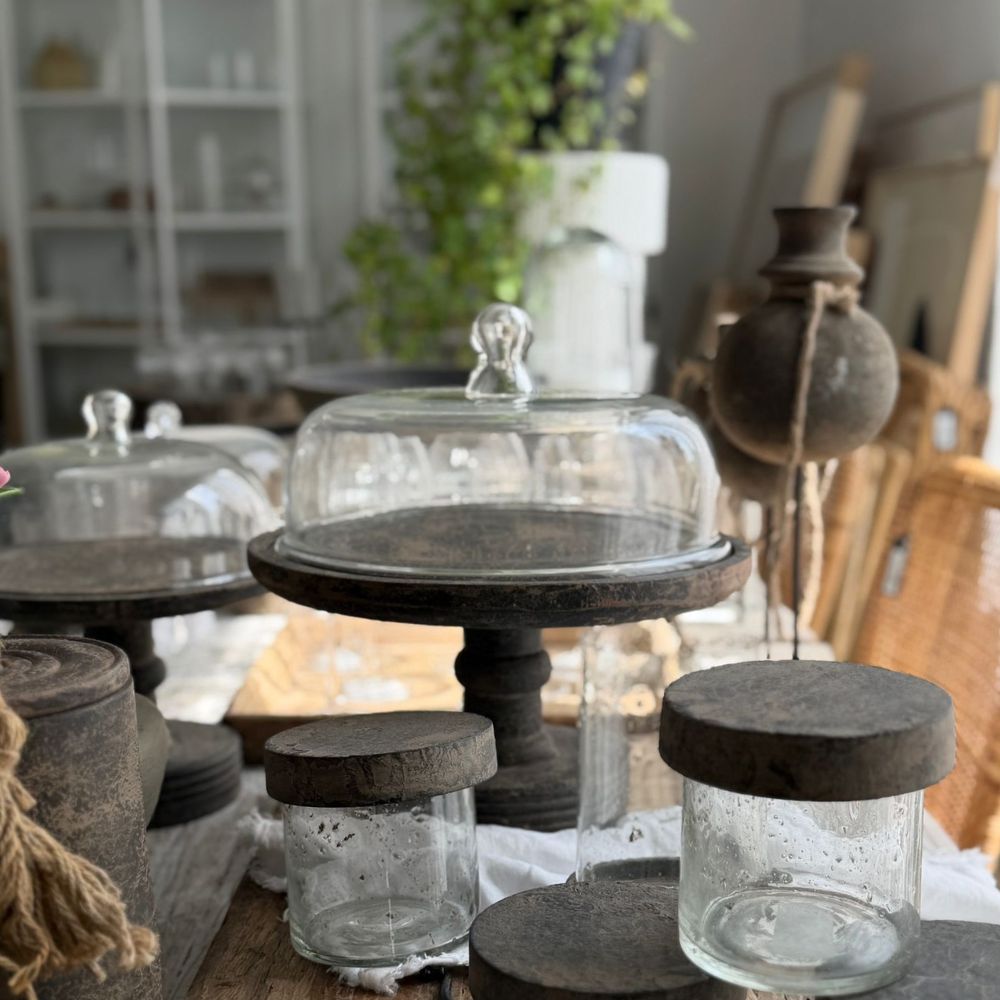 פעמון עוגה על רגל עם מכסה זכוכית מבית Fine Lab Home מונח על שולחן עץ, כשברקע נראים צמחים ומדפים.