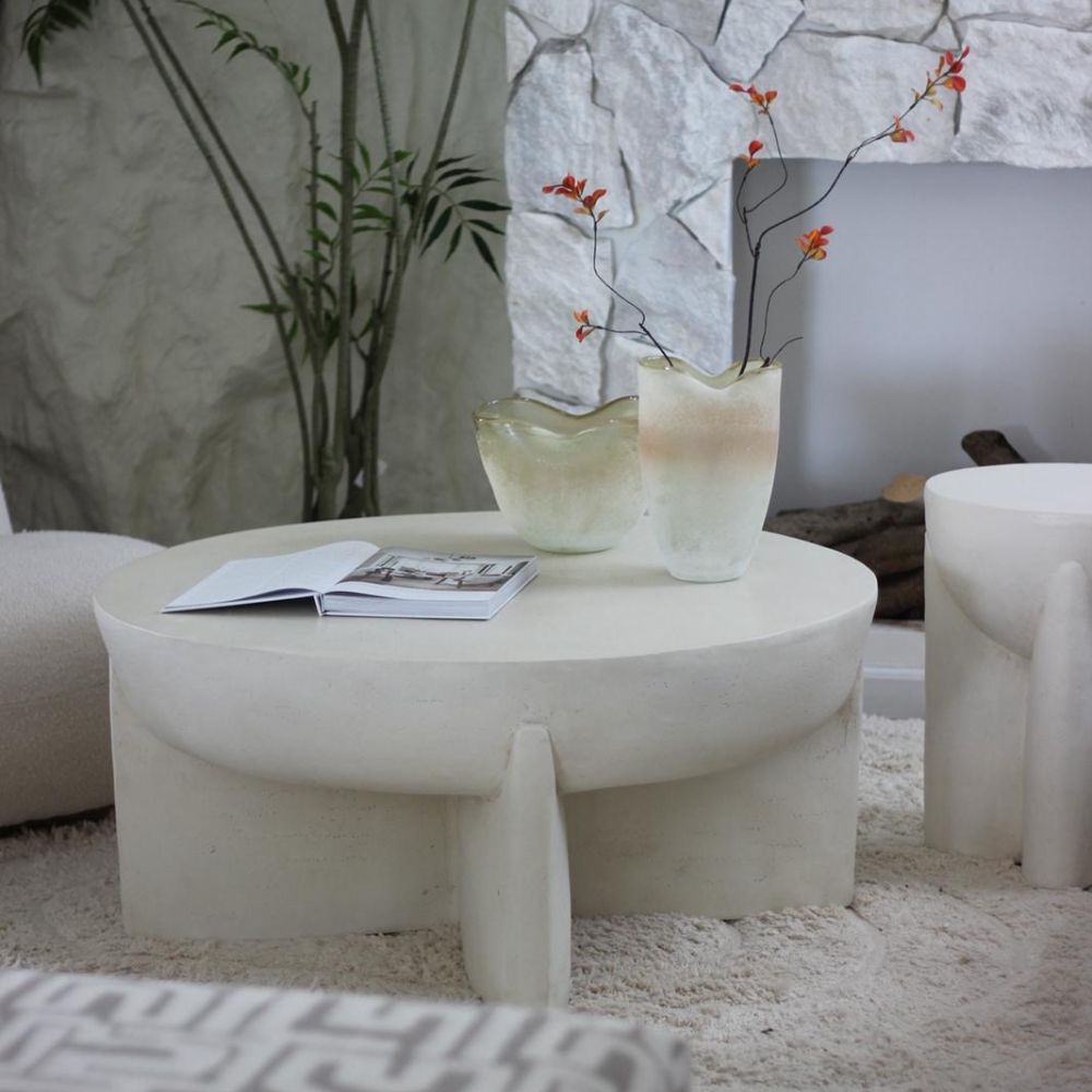 השולחן הסלון העגול דמוי אבן מבית Fine Lab Home עם שני אגרטלי זכוכית מלאים בפרחי כתום וספר פתוח, מונח על שטיח בהיר ומשדר מראה מודרני ומינימליסטי לסלון.